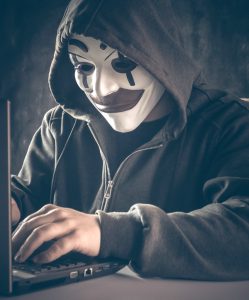 male hacker with anonymous clown mask in hoodie on laptop